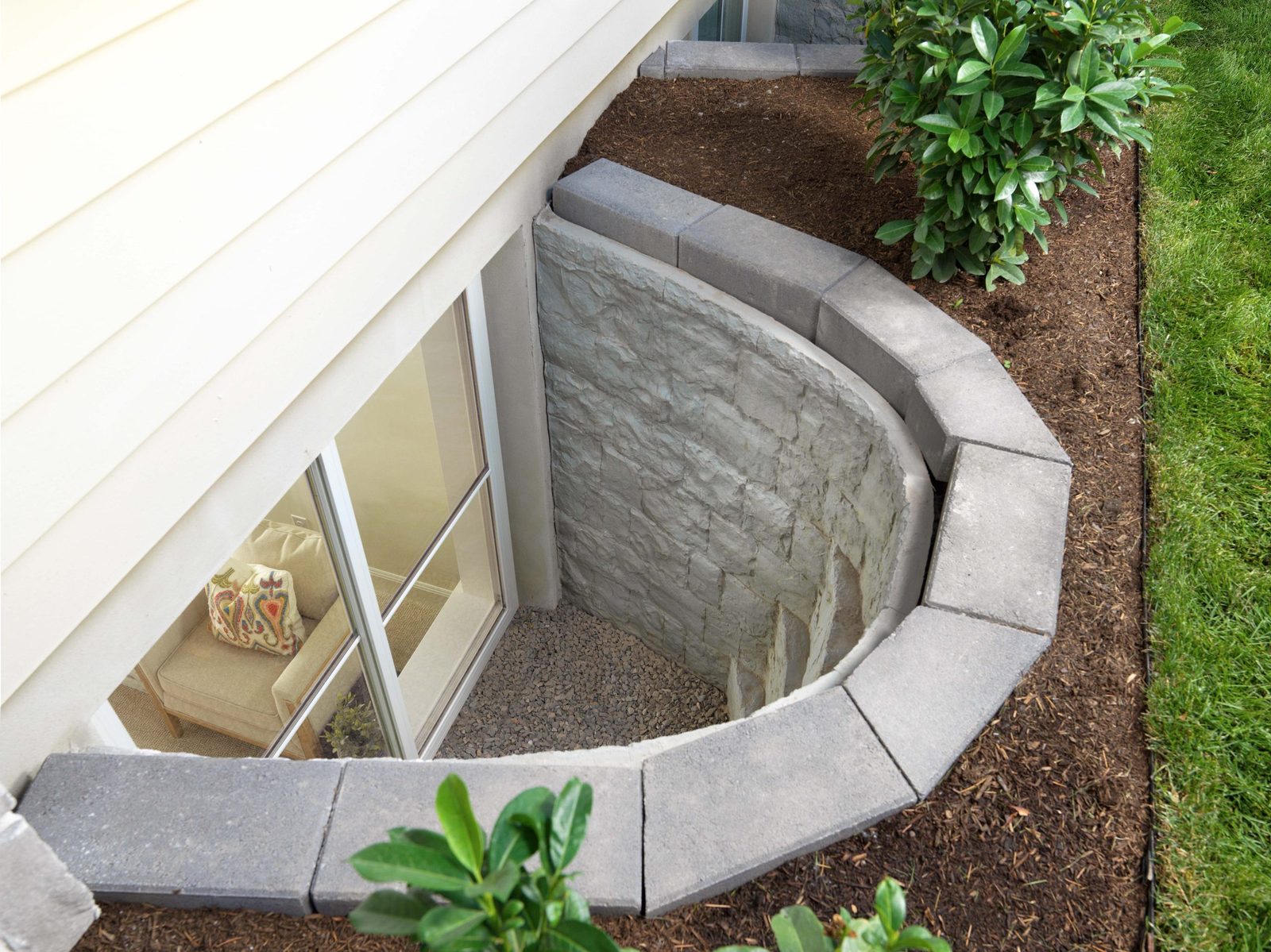 Outside view of a gray stone-look window well with built-in steps and a gravel floor beside a house